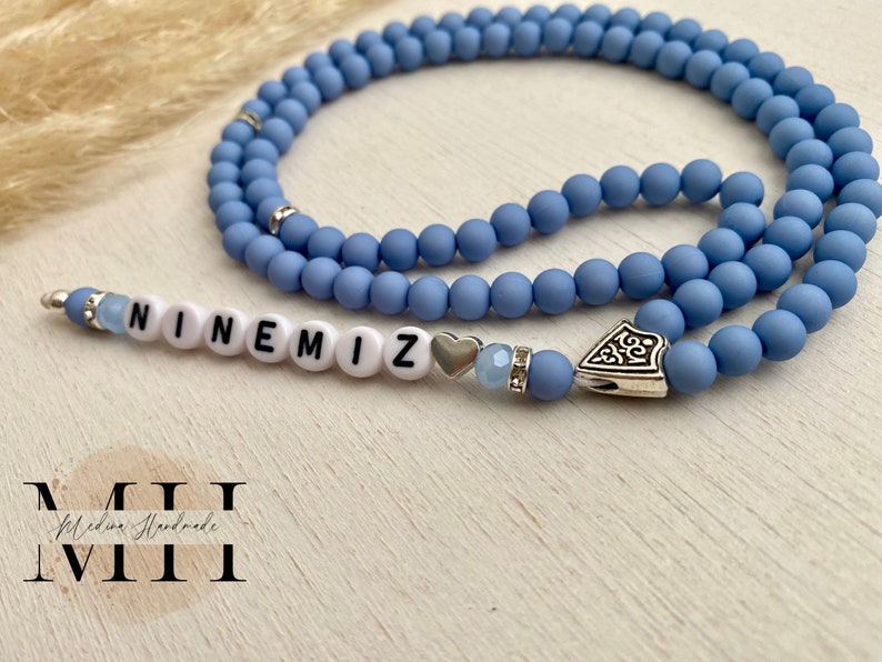 Op de afbeelding: Een lichtblauwe kralenketting met het woord "NINEMIZ" gespeld in witte letterkralen. De ketting heeft zilverkleurige accenten, waaronder een hartvormige bedel en een decoratieve hanger.