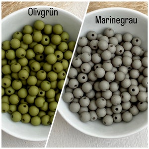 Op de afbeelding: Twee witte kommen gevuld met ronde kralen. E&eacute;n kom bevat olijfgroene kralen en de andere marinegrijze kralen. De tekst "Olivgr&uuml;n" staat boven de olijfgroene kralen en "Marinegrau" boven de grijze kralen.