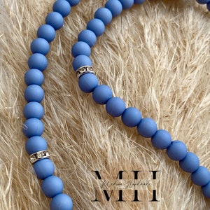 Op de afbeelding: Een close-up van een lichtblauwe kralenketting met zilveren strass accenten. De ketting is gemaakt van ronde kralen en wordt getoond op een beige, getextureerd oppervlak. De ketting heeft de tekst "MII" in witte letters.