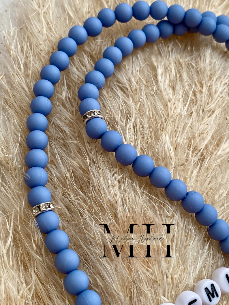 Op de afbeelding: Een close-up van een lichtblauwe kralenketting met zilveren strass accenten. De ketting is gemaakt van ronde kralen en wordt getoond op een beige, getextureerd oppervlak. De ketting heeft de tekst "MII" in witte letters.