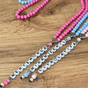 Op de afbeelding: Close-up van drie kralenkettingen in roze, blauw en lichtroze. Elke ketting heeft witte letterkralen die een naam spellen. De kralen zijn geregen op een zilveren ketting met decoratieve eindkappen. De achtergrond is een houten oppervlak.