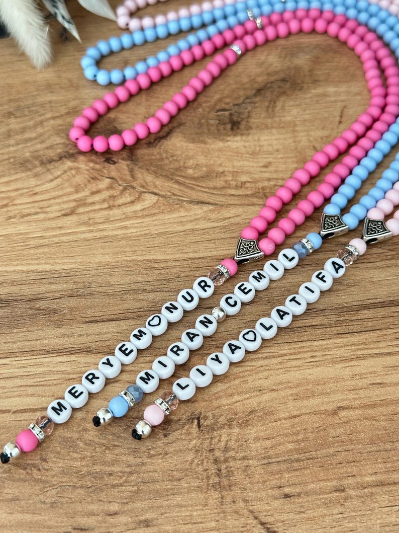 Op de afbeelding: Close-up van drie kralenkettingen in roze, blauw en lichtroze. Elke ketting heeft witte letterkralen die een naam spellen. De kralen zijn geregen op een zilveren ketting met decoratieve eindkappen. De achtergrond is een houten oppervlak.