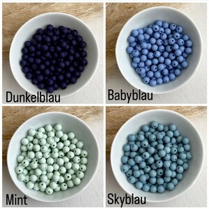 Op de afbeelding: Vier kleine witte kommen, elk gevuld met ronde kralen in verschillende tinten blauw en groen. De kleuren zijn donkerblauw, lichtblauw, mintgroen en een gedempt blauwgrijs. Tekstlabels identificeren elke kleur.