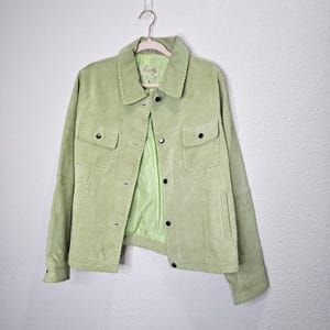 Peut inclure: Veste en daim vert clair avec un col et une fermeture boutonnée. La veste comporte deux poches de poitrine et des coutures décoratives. La doublure intérieure est d'un vert plus clair. La veste est suspendue à un cintre rose.