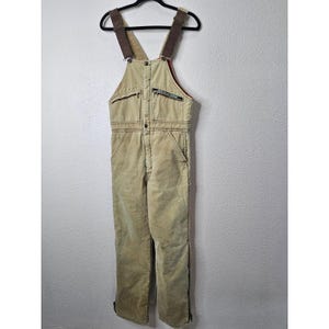May include: Tan canvas dungarees featuring brown adjustable shoulder straps, a front bib pocket with a zip closure, and a small rectangular patch with text. The garment includes side pockets, a front button placket, and full-length leg zips.