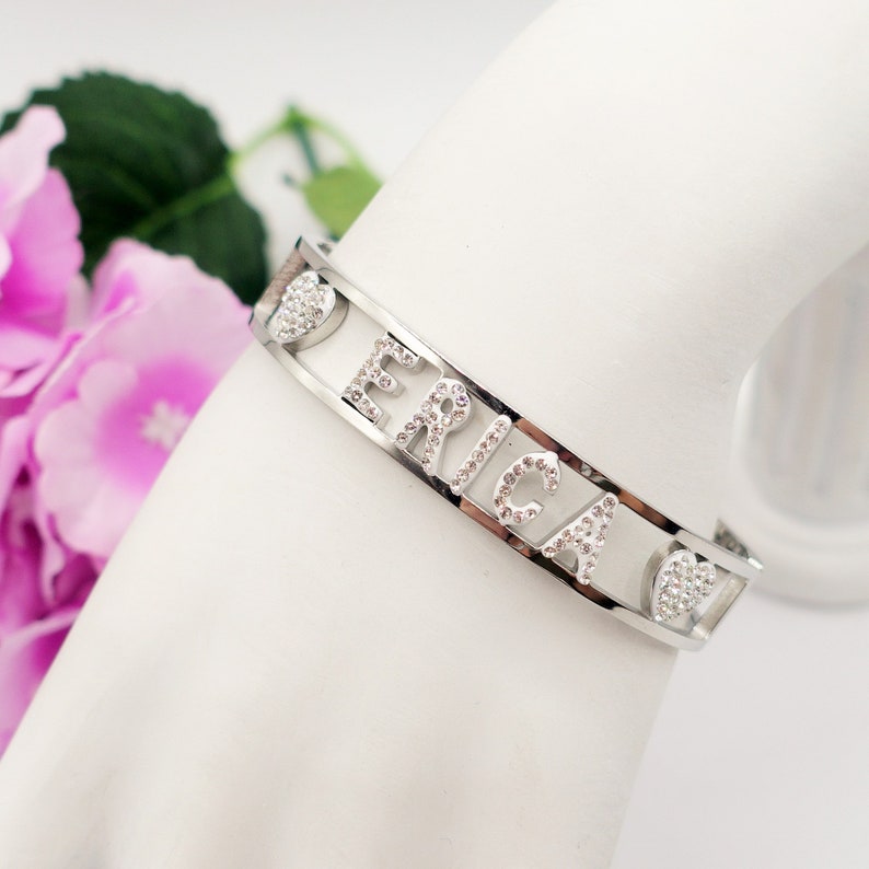 May include: Silver bangle bracelet with the name "ERICA" spelled out in clear rhinestones. The bracelet has two heart-shaped cutouts on either side of the name, also decorated with rhinestones.