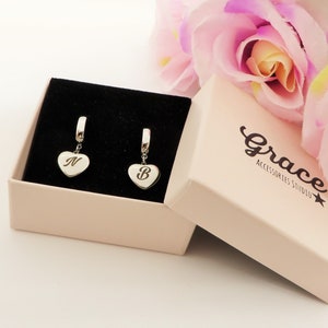 May include: A pair of silver heart-shaped earrings, each with a letter engraved on the front, displayed in an open pink box. The box is lined with black velvet and has the words "Grace Accessories Studio" printed on the side. Pink flowers are in the background.