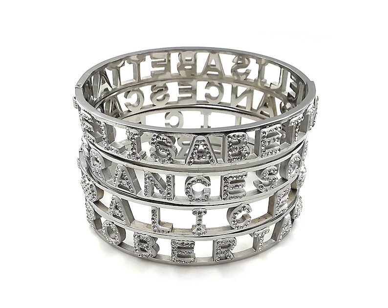 May include: Four silver bracelets stacked on top of each other. Each bracelet is made of metal and has a different name spelled out in the metal. The names are "Elizabeth", "Orange", "Alice", and "Robert".