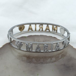 May include: Silver bangle bracelet with the name "MARIA" spelled out in silver letters with clear rhinestones. The bracelet has two heart charms, one with a gold heart and the other with clear rhinestones.