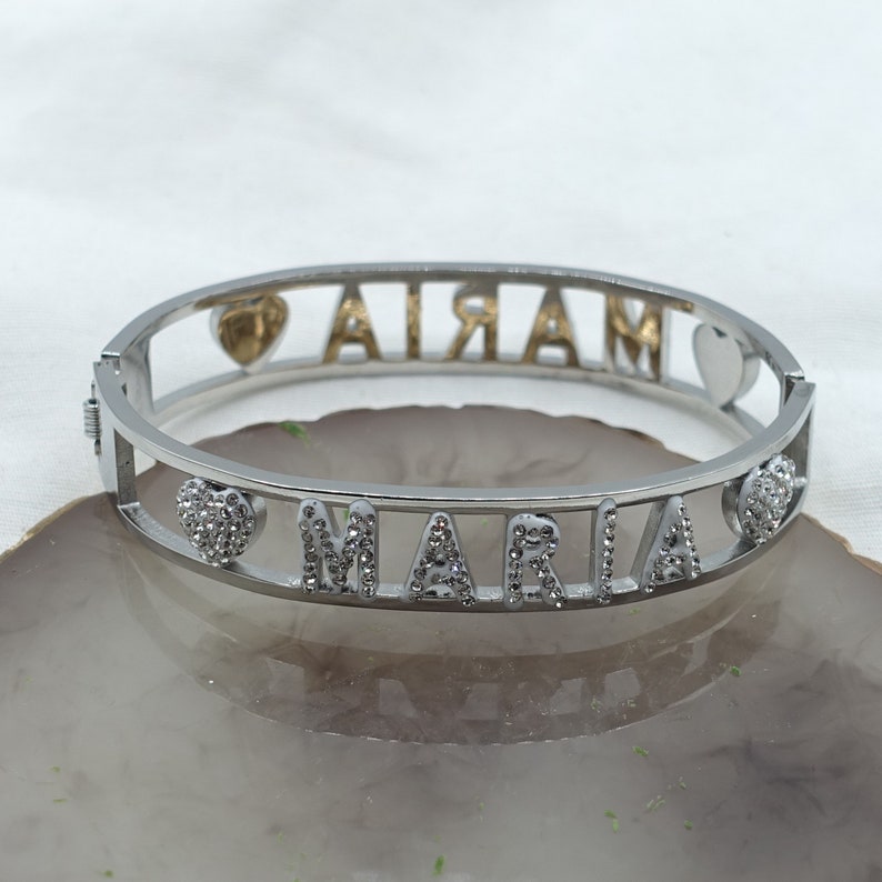 May include: Silver bangle bracelet with the name "MARIA" spelled out in silver letters with clear rhinestones. The bracelet has two heart charms, one with a gold heart and the other with clear rhinestones.
