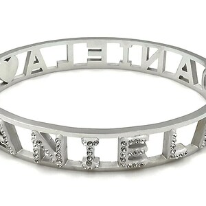 May include: Silver bangle bracelet with the word "VALENTINA" spelled out in cut-out letters. The letters are outlined with small, clear rhinestones.