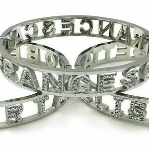 May include: Three silver bangle bracelets with the words "ORANGE", "ROBERT", and "ELISABETH" spelled out in raised letters. Each bracelet is adorned with small, clear rhinestones.