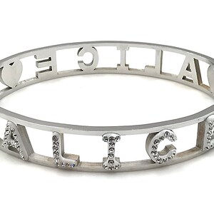 May include: A silver bangle bracelet with the word "ALICE" spelled out in cut-out letters. Each letter is decorated with small, clear rhinestones. The bracelet has two heart charms on either side of the word.