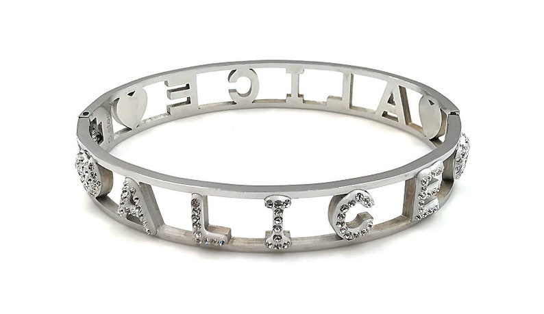 May include: A silver bangle bracelet with the word "ALICE" spelled out in cut-out letters. Each letter is decorated with small, clear rhinestones. The bracelet has two heart charms on either side of the word.