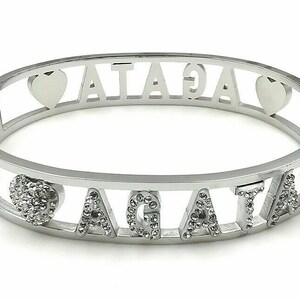 May include: Silver bangle bracelet with the name "AGATHA" spelled out in the center. The letters are decorated with small clear rhinestones. There are two heart-shaped charms on the bracelet, one on each side of the name. One heart is plain silver, and the other is decorated with rhinestones.