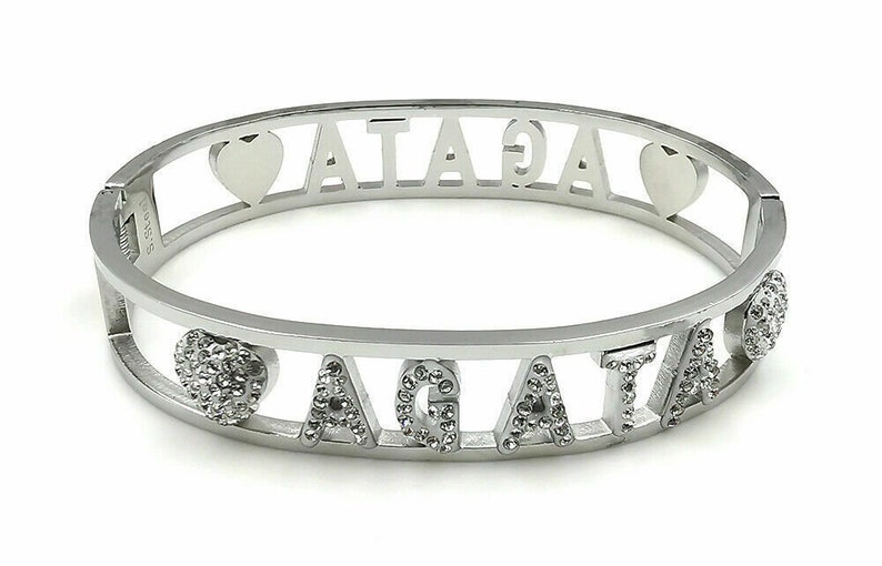 May include: Silver bangle bracelet with the name "AGATHA" spelled out in the center. The letters are decorated with small clear rhinestones. There are two heart-shaped charms on the bracelet, one on each side of the name. One heart is plain silver, and the other is decorated with rhinestones.