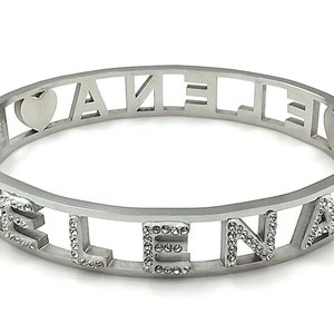 May include: Silver bangle bracelet with the name "ELENA" spelled out in cut-out letters with small rhinestones around each letter. The bracelet has a heart charm on each side.