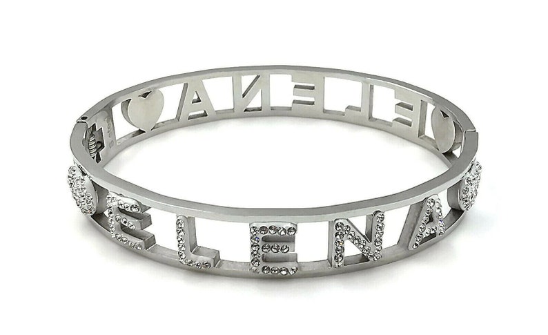 May include: Silver bangle bracelet with the name "ELENA" spelled out in cut-out letters with small rhinestones around each letter. The bracelet has a heart charm on each side.