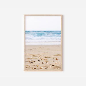 Op de afbeelding: Een ingelijste prent van een strandscène. De afbeelding toont een zandstrand op de voorgrond met kleine kiezels en schelpen. De oceaan is op de achtergrond, met blauw water en witte golven onder een heldere hemel. De lijst is licht van kleur.