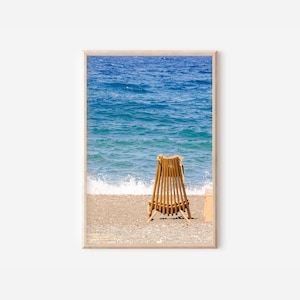 Op de afbeelding: Een ingelijste prent met een strandscène. Een houten strandstoel staat op een kiezelstrand, met uitzicht op de zee. Het water is helderblauw en de lucht is helder. De stoel is gemaakt van verticale houten latten.