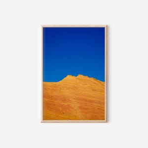 May include: A framed art print featuring a vibrant landscape. The image showcases a striking contrast between a clear, bright blue sky and a textured, orange-yellow rock formation. The artwork is presented in a natural wood frame.
