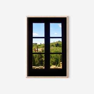 Op de afbeelding: Een ingelijste print met een uitzicht door een zwart raam. Het raam is verdeeld in zes ruiten en biedt een blik op een groene omgeving onder een helderblauwe lucht. De scène omvat bomen, gebouwen en glooiende heuvels.