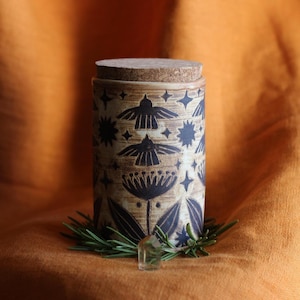 May include: A cylindrical ceramic jar with a cork lid, featuring a dark brown sgraffito pattern of birds, stars, and botanical elements on a light tan clay body. The jar is surrounded by a small ring of green rosemary sprigs and a clear crystal.