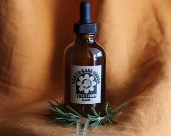 May include: An amber glass dropper bottle with a black cap and a white label featuring a flower graphic, a figure, and the text Earth-Babe-Herbs Tremella Elixir. A sprig of green rosemary and a small clear crystal are placed at the base of the bottle.
