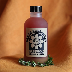May include: A frosted glass bottle filled with a reddish liquid, topped with a black cap. The label features a black flower graphic with a central figure and the text: EARTH-BABE-HERBS FIRE CIDER VINEGAR + HONEY. A sprig of green rosemary encircles the base.