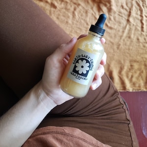 May include: A clear glass bottle with a dropper top filled with a light yellow liquid. The bottle has a white label with black text that reads "Earth Baby Things Shatavari Elixir".