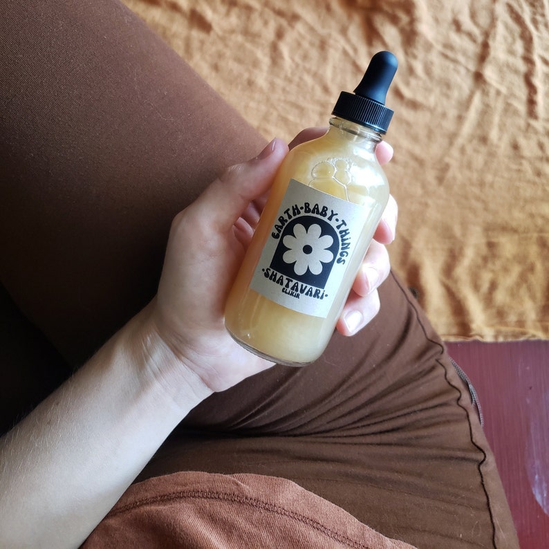 May include: A clear glass bottle with a dropper top filled with a light yellow liquid. The bottle has a white label with black text that reads "Earth Baby Things Shatavari Elixir".