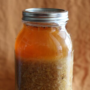May include: A glass jar with a silver lid filled with a light brown liquid and a layer of white, translucent solids at the bottom.
