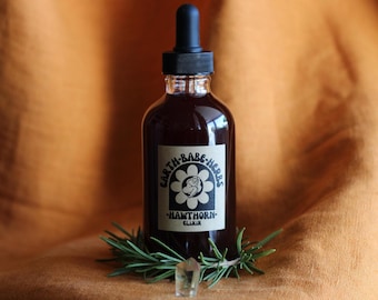 Puede incluir: Un frasco cuentagotas de vidrio transparente lleno de líquido oscuro, con una etiqueta color canela con el texto Earth-Babe-Herbs Hawthorn Elixir y un gráfico de una persona dentro de una flor. Una ramita de romero verde y un pequeño cristal claro en la base.