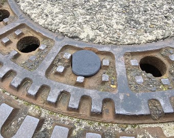 Puede incluir: Primer plano de una tapa de alcantarilla de metal oxidado con un tapón de goma negro en el centro.