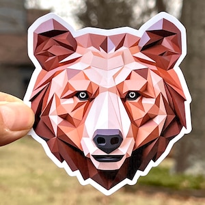 May include: A sticker featuring a geometric bear head design. The bear's face is composed of various shades of brown and tan polygons, with black eyes and a black nose. The sticker has a white border.