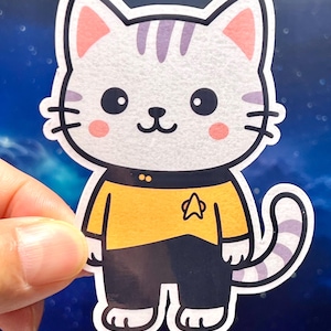 May include: A sticker featuring a cartoon cat with a white body, purple stripes, and pink ears. The cat wears a yellow uniform with a black collar and pants. A star emblem is on the chest. The cat has a smiling face with pink cheeks.