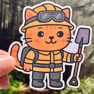 May include: A sticker featuring an orange cartoon cat dressed as a firefighter. The cat wears a yellow helmet with goggles, a yellow uniform, and gray pants. It holds a small shovel. The sticker has a white border and is held against a blurred forest background.