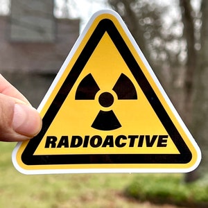 May include: A yellow triangular sticker with a black border and a black radiation symbol. The word "RADIOACTIVE" is printed in black, centered below the symbol. The sticker is held against a blurred outdoor background.