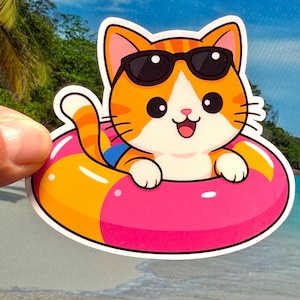 May include: A die-cut sticker featuring an orange and white tabby cat wearing black sunglasses while floating in a pink and yellow inflatable pool ring. The cat has a happy expression with an open mouth and is wearing blue swimming trunks.