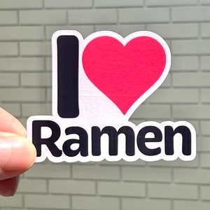K&ouml;nnte beinhalten: Ein Aufkleber mit den Worten "I &hearts; Ramen". Das "I" ist ein schwarzer Gro&szlig;buchstabe, das Herz ist rot und das Wort "Ramen" ist in schwarzen, fetten Buchstaben geschrieben. Der Aufkleber wird von den Fingern einer Person gehalten.