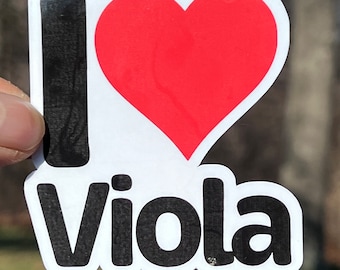 Puede incluir: Una pegatina blanca con el texto "Me encanta la viola" en letras negras. Un corazón rojo está entre la "M" y la "encanta".