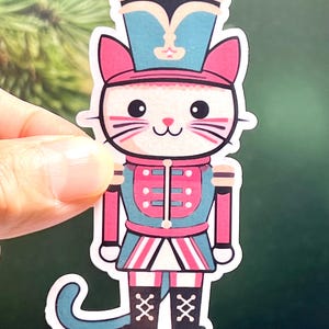 May include: A white cat dressed as a Christmas nutcracker. The cat is wearing a pink and blue hat, a red and blue uniform, and black boots. The cat has a blue tail.