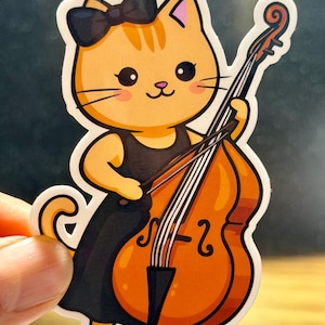 May include: A sticker featuring an orange tabby cat playing a double bass. The cat wears a black dress and a black bow. The double bass is a warm orange color with black strings. The sticker has a white border.