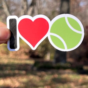 May include: A sticker with the words "I ❤️ Tennis". The "I" is black, the heart is red, and the tennis ball is green with white accents. The sticker is held up against a blurred outdoor background.