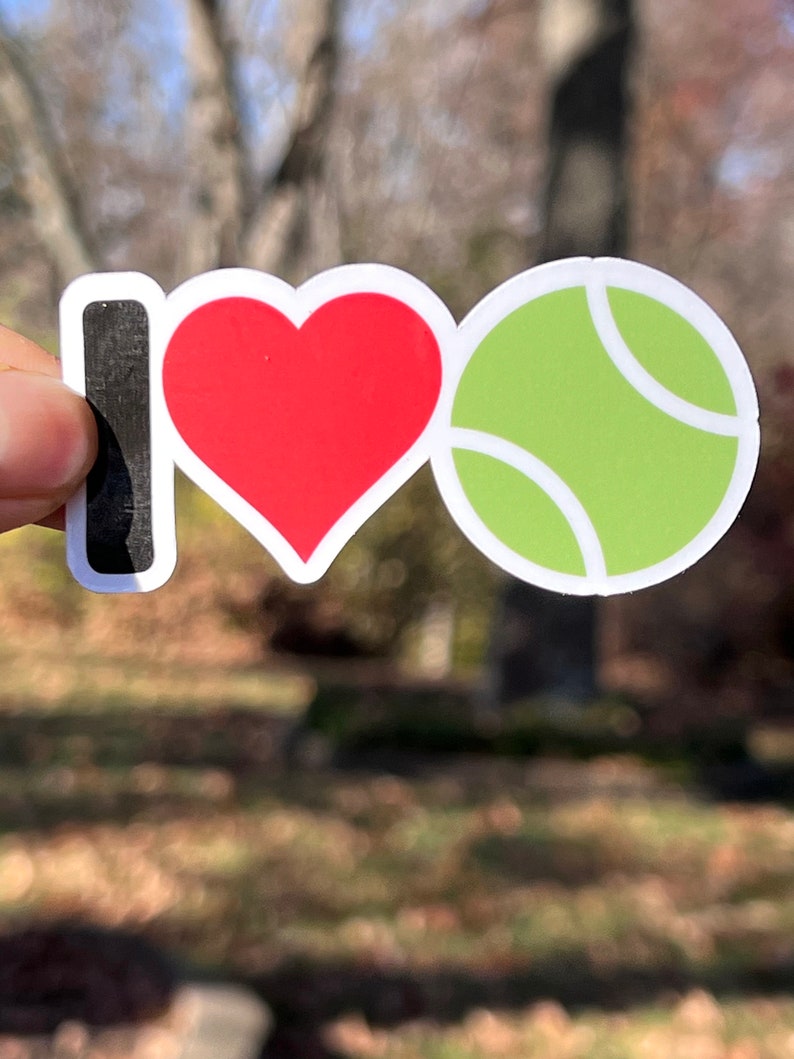 May include: A sticker with the words "I ❤️ Tennis". The "I" is black, the heart is red, and the tennis ball is green with white accents. The sticker is held up against a blurred outdoor background.