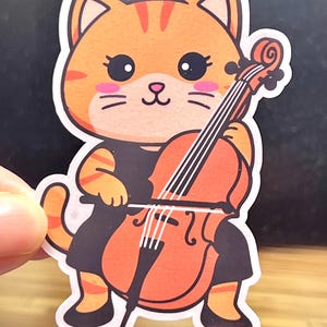 May include: A sticker featuring a cartoon cat playing a cello. The cat is orange with black stripes and wears a black dress. The cello is orange and brown with white strings. The sticker has a white border.