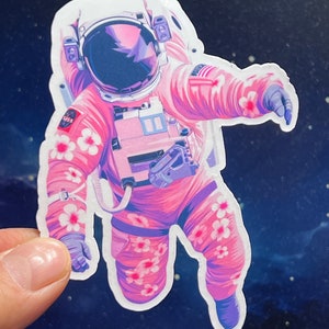 May include: A pink astronaut with white flowers on their suit floats in space against a dark blue background with white stars.