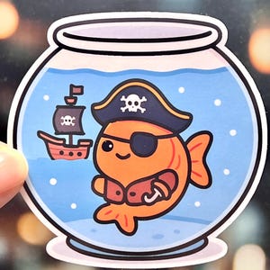 May include: A sticker featuring an orange goldfish in a fishbowl, dressed as a pirate. The fish wears a black pirate hat with a skull and crossbones and an eye patch. A small pirate ship is also included in the design.
