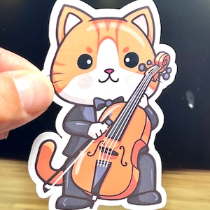 May include: A die-cut sticker featuring an orange and white tabby cat wearing a black tuxedo and bow tie. The cat is standing upright and playing a large, orange-colored cello with a bow, depicted in a cartoon illustration style with bold outlines.