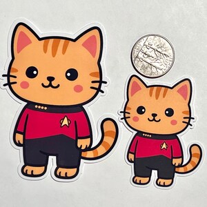 May include: Two orange tabby cat stickers, one large and one small, wearing red and black uniforms with a gold insignia. A US quarter is included for size comparison. The cats have large black eyes and pink cheeks.